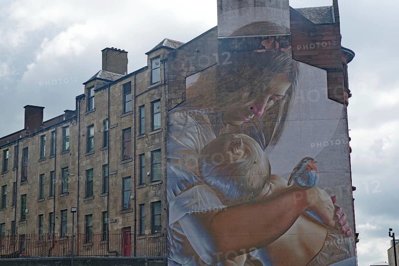 A mural shows a woman holding a baby and a bird in her arms. A building can be seen in the background, Mural Trail, Glasgow, Scotland, Great Britain