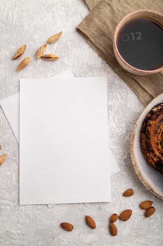 White paper invitation card, mockup with almonds, bun, cup of coffee, linen textile on gray concrete background. Blank, flat lay, top view, still life, copy space