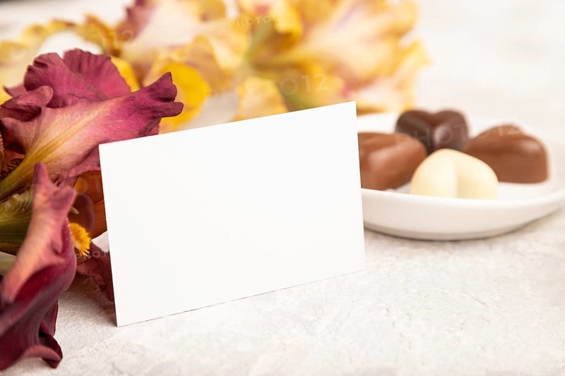 White business card with chocolate candies and iris flowers on gray concrete background. side view, copy space, still life. Breakfast, morning, spring concept