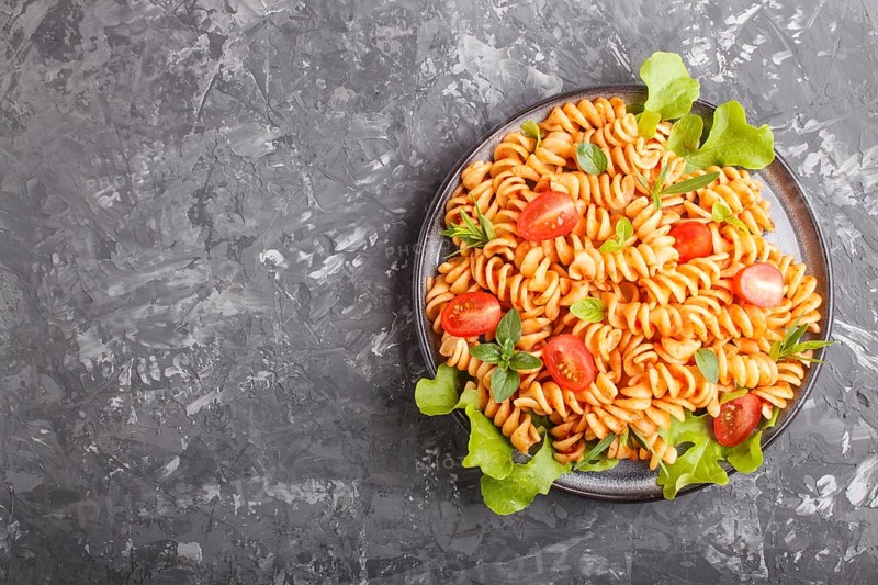 Fusilli pasta with tomato sauce, cherry tomatoes, lettuce and herbs on a black concrete background. top view, flat lay, copy space