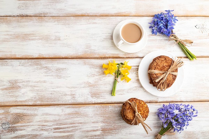 Oatmeal cookies with spring snowdrop flowers bluebells, narcissus and cup of coffee on white wooden background. top view, flat lay, copy space, still life. Breakfast, morning, spring concept