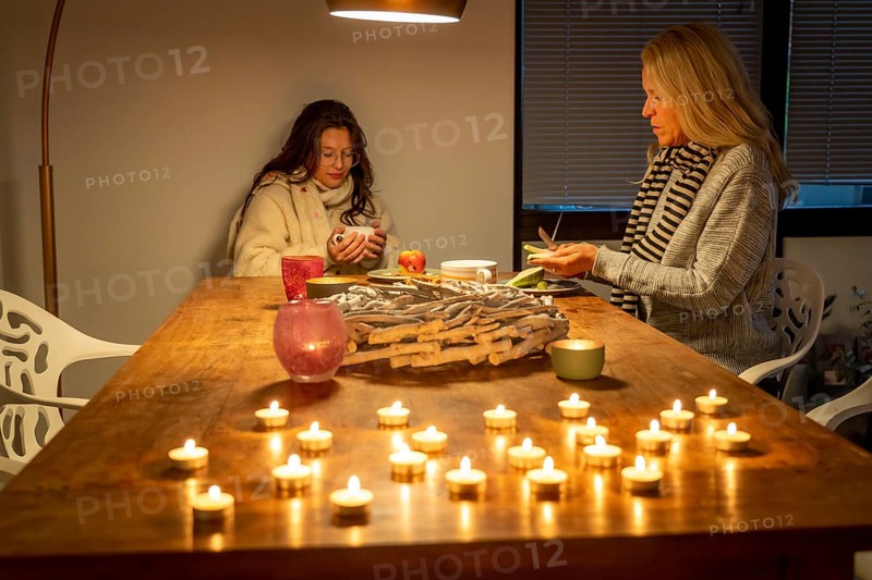Symbolic image energy saving, cold winter, energy crisis, cold flat, mother and daughter dressed warmly at dinner, only one lamp on, candlelight, heating on to a minimum