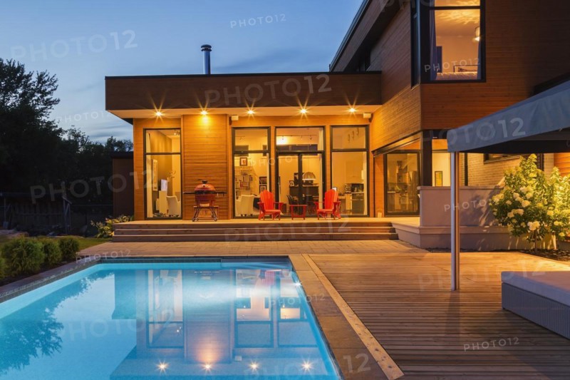 Rear of modern cube style home with stained horizontal wood cladding, in-ground swimming pool and wooden deck with gazebo canopy at dusk, Quebec, Canada, North America