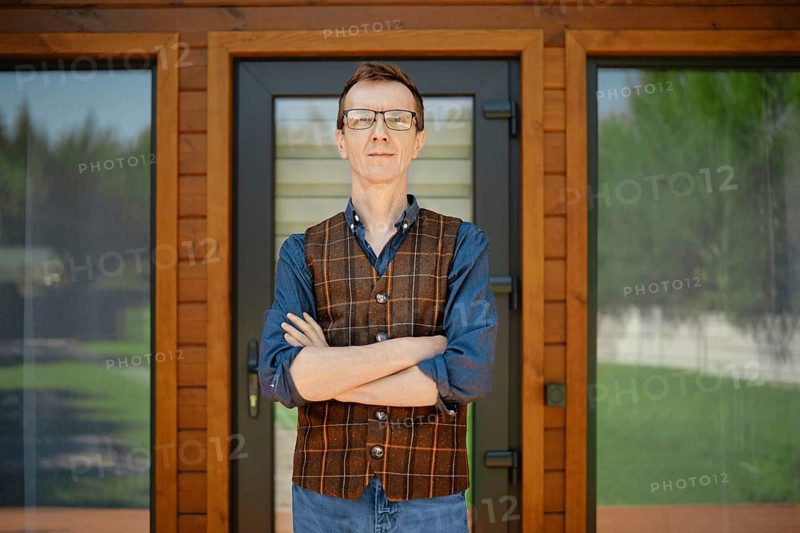 Middle-aged man in blue shirt and brown vest stands confidently in front of modern wooden house doors