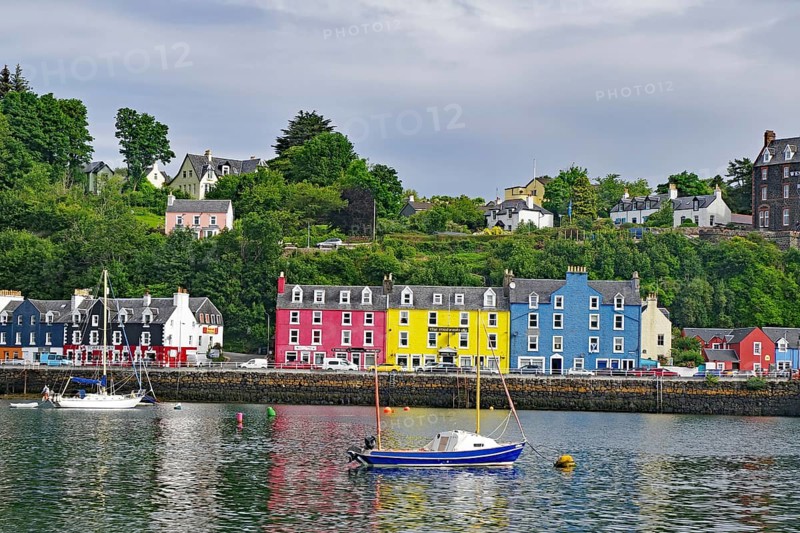 Houses with colourful facades along a harbour, boats in the foreground and a green hill in the background under a blue sky with clouds, Tobermory, Mull, Inner Hebrides, Scotland, Great Britain