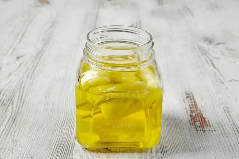 A square glass jar on a white wooden table. The jar is filled with a pickled yellow Chilli peppers