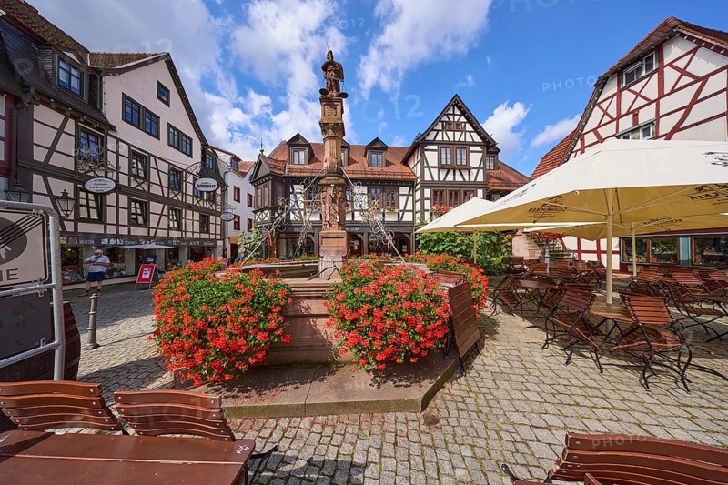 Market fountain with geraniums and outdoor area of a restaurant under a blue sky with cumulus clouds on the market square in Michelstadt, Odenwald, Odenwaldkreis, Hesse, Germany, Europe