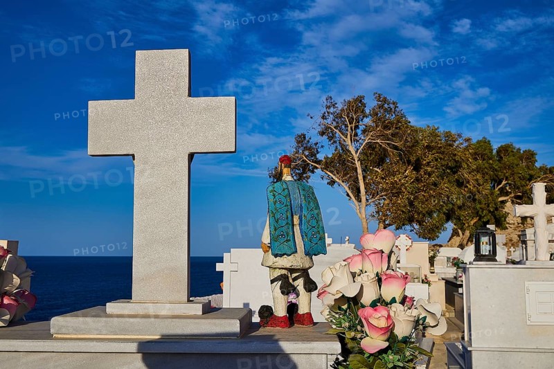 A cemetery by the sea with gravestones and crosses. In the foreground are flowers and a figure with a colourful cloak, Agios Nikolaos church, cemetery, Pigadia, town and harbour, Pigadia Bay, main town, Karpathos, Dodecanese, Greek Islands, Greece, Europe