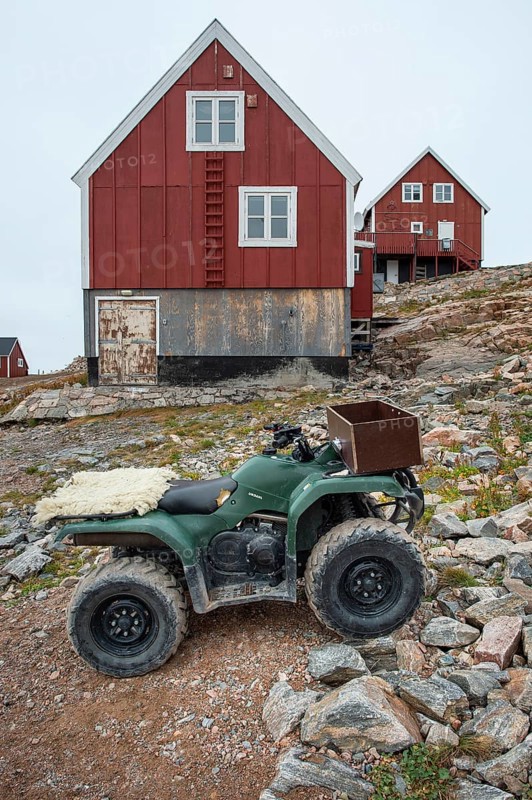 A green quad bike or ATV stands in front of red, wood-clad houses on a rocky hill, remote Arctic Inuit settlement Ittoqqortoormiit, Scoresbysund or Scoresby Sund or Greenlandic Kangertittivaq, East Greenland, Greenland, North America