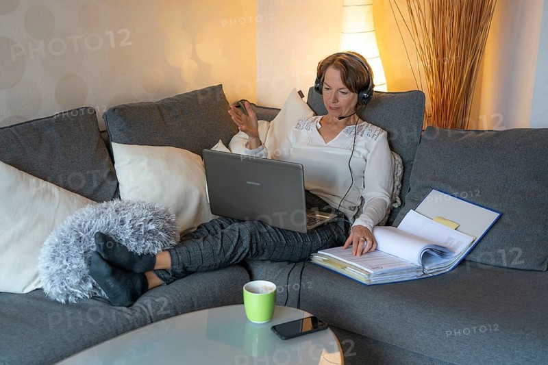 Woman, mid-50s, works from home, with laptop and communicates with colleagues via headset, home office, on the sofa