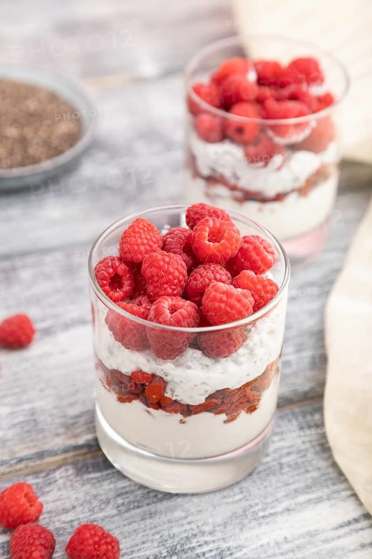 Yogurt with raspberry, goji berries and chia seeds in glass on gray wooden background and linen textile. Side view, close up, selective focus