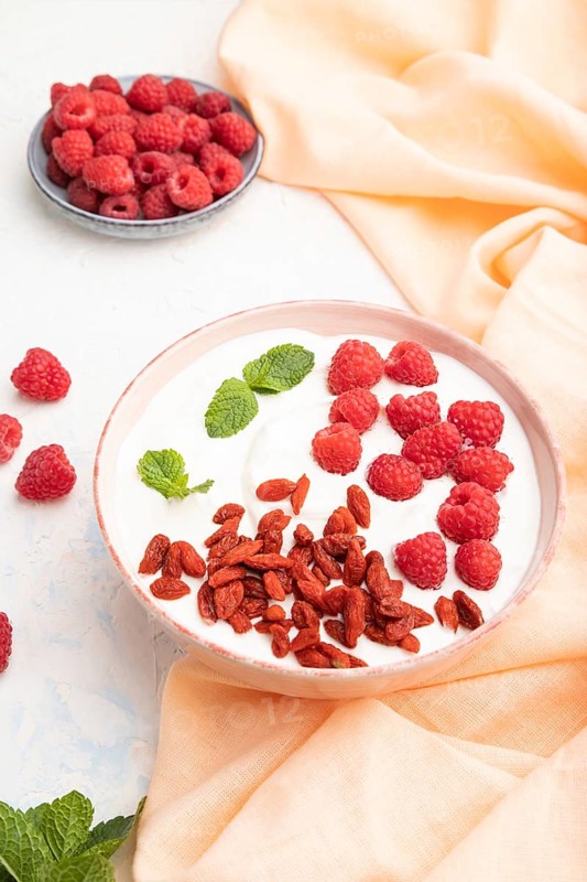 Yogurt with raspberry and goji berries in ceramic bowl on white concrete background and orange linen textile. Side view, close up