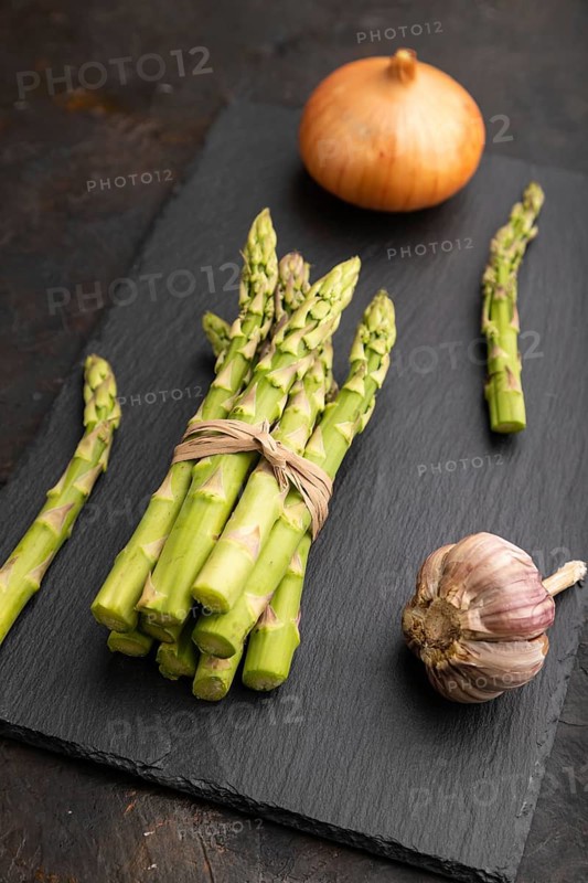 Bunch of fresh green asparagus, garlic, onion on slate board on black concrete background. Side view, close up. harvest, healthy, vegan food, concept