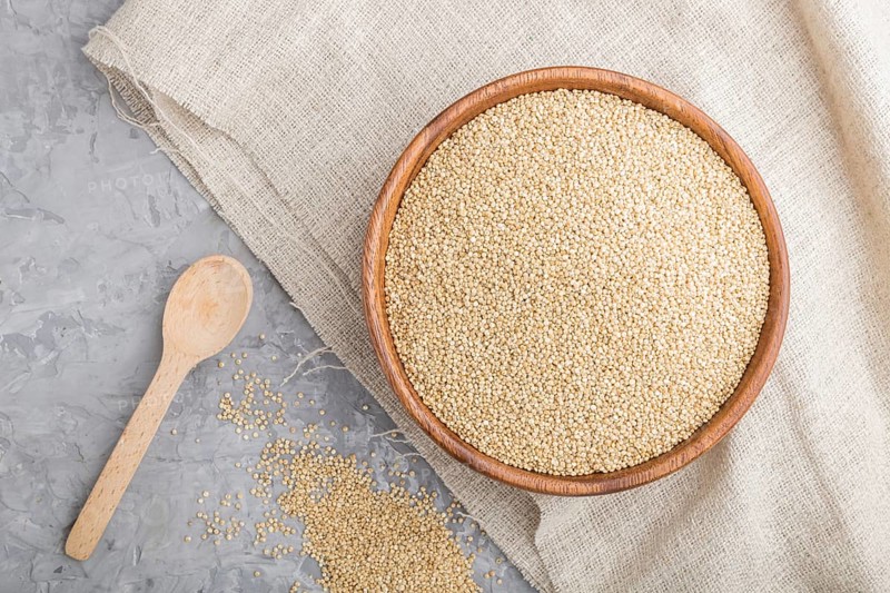 Wooden bowl with raw white quinoa seeds and wooden spoon on a gray concrete background and linen textile. Top view, flat lay, close up