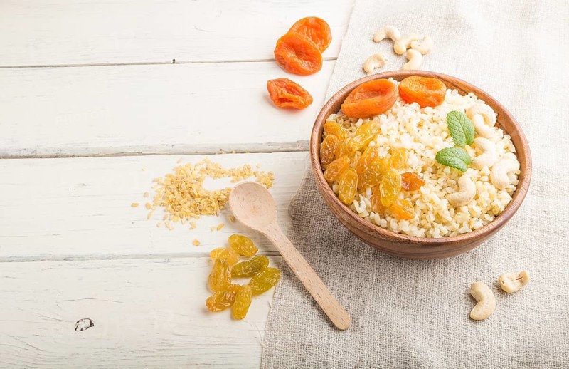 Bulgur porridge with dried apricots, raisins and cashew in wooden bowl on a white wooden background and linen textile. Side view, copy space. Turkish traditional cuisine