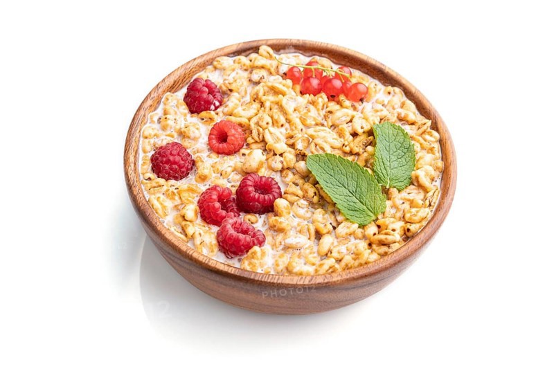Wheat flakes porridge with milk, raspberry and currant in wooden bowl isolated on white background. Side view, close up