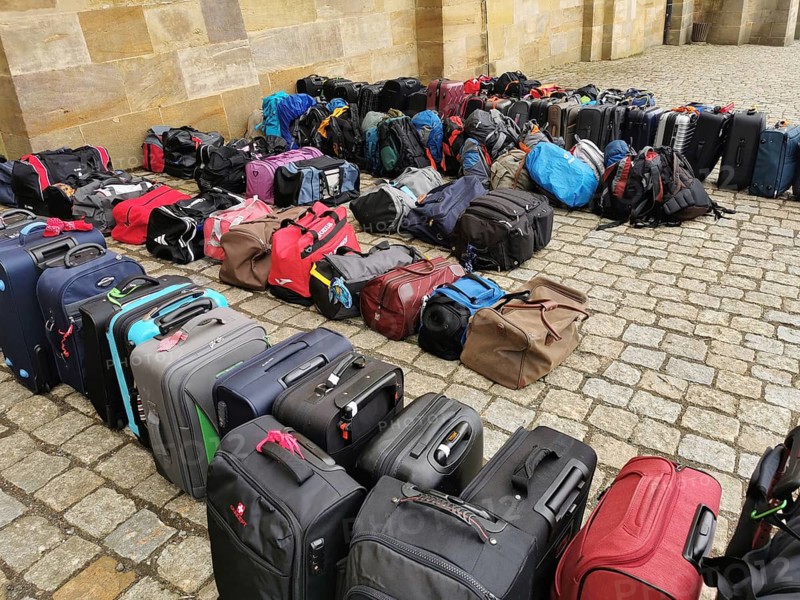 The luggage of a group of travellers stands in rows on the pavement, outside, suitcases and bags