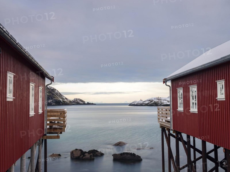 View between two traditional red wooden houses across the fjord to the mountains of the mainland, Nusfjord, Flakstadøya, Lofoten, Norway, Europe