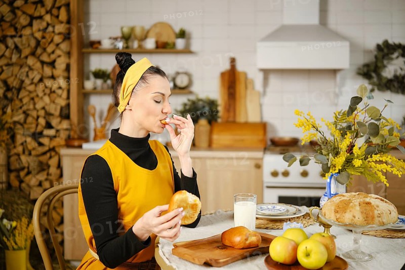Woman eating an apple bun breaking off pieces of soft dough. She is sitting in a wel-lit country-style kitchen with wooden decor