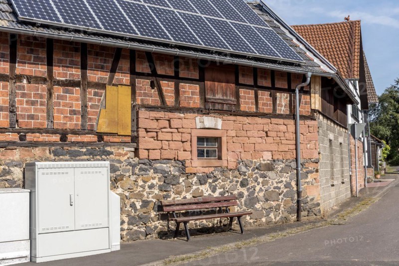 Historic farmhouse with photovoltaic system, solar panels on the roof, house wall with differently pieced masonry made of brick, sandstone, fieldstone and half-timbering, in front of it electricity distribution box and bench, Nidda, Hesse, Germany, Europe