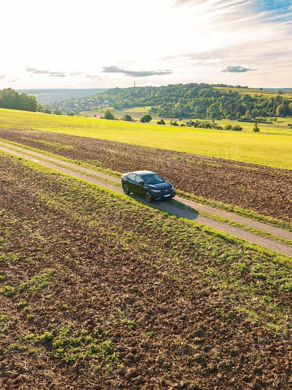 Black car driving on a gravelled country lane through a wide, rural landscape under a cloudy sky, Carsharing, VW ID5, Calw, Black Forest, Germany, Europe