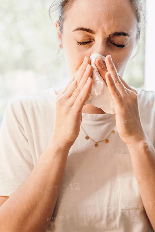 Woman in bright room uses paper towel, focus is on facial expression of discomfort, 'symbolic image' of a cold