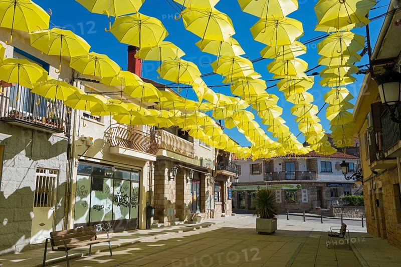 Sunny alley with yellow hanging umbrellas as decoration, surrounded by various houses under a clear blue sky, historic centreLos Molinos, Cuenca del Guadarrama, Madrid, Spain, Europe