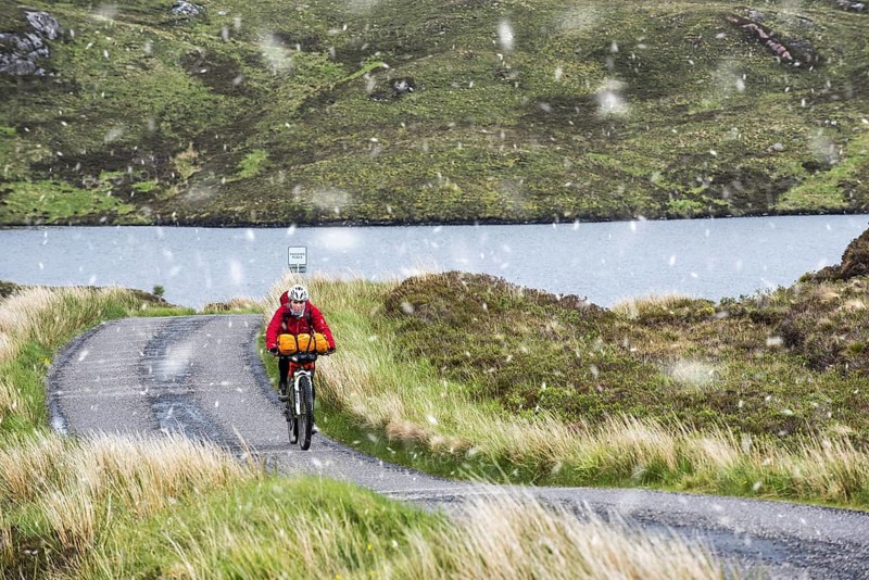 Lonely biker cycling through the Scottish Highlands on heavily laden touring bicycle along single track road during sleet in spring, Scotland, UK