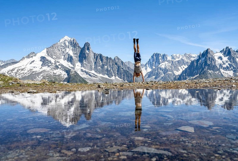 Young man doing a handstand in front of a mountain landscape, reflection in a small mountain lake, mountain peaks Aiguille Verte, Grandes Jorasses and Mont Blanc, mountain panorama with Mont Blanc massif, Aiguille Rouge, Chamonix-Mont-Blanc, Haute-Savoie, France, Europe