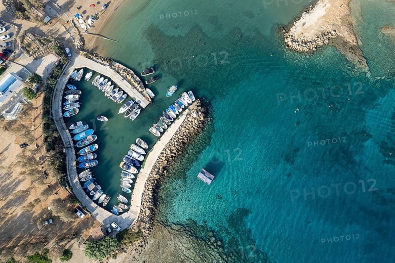 Aerial drone view of fishing harbour and sandy beach. Agia Triada people swimming and fishing boats moored at the harbor. Protaras Paralimni Cyprus