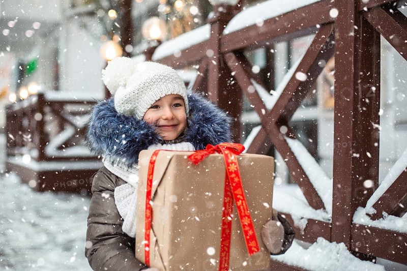 Portrait of joyful girl with a gift box for Christmas on a city street in winter with snow on a festive market with decorations and fairy lights. Warm clothes, knitted hat, scarf and fur. New year