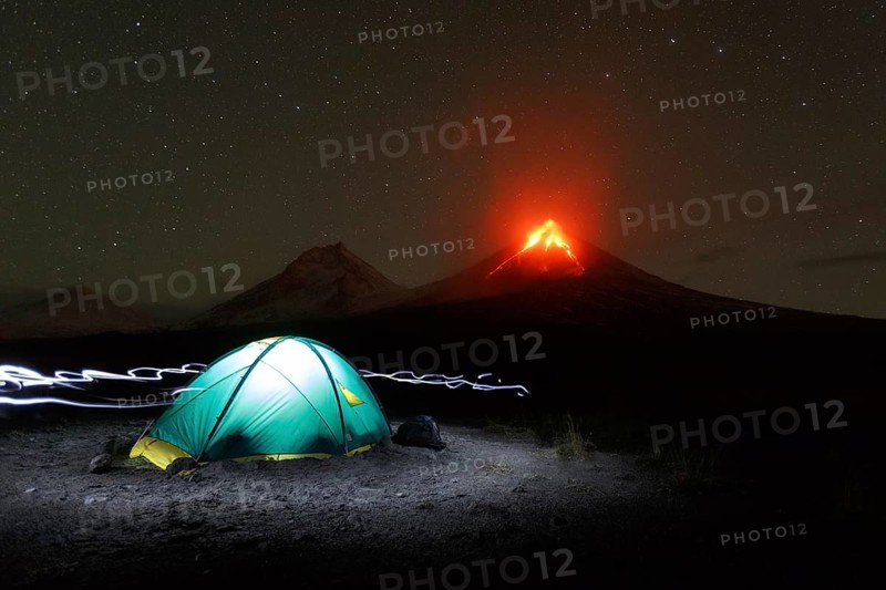Night mountain landscape of Kamchatka Peninsula: illuminated tourist camping on background eruption volcano, light from headlights around tent. Russian Far East, Kamchatka Region, Klyuchevskoy Volcano