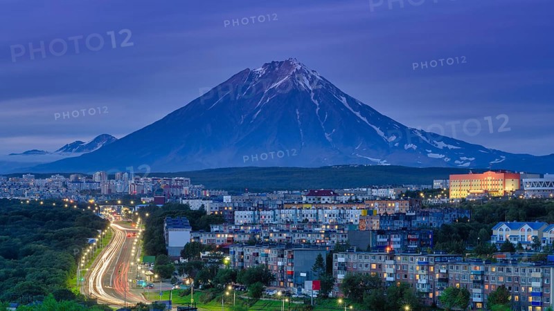 Panorama of night city of Petropavlovsk-Kamchatsky on background cone of volcano, urban development at twilight, backlit city road with driving car lights