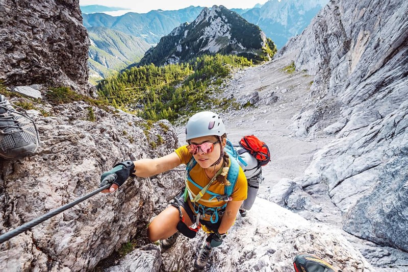 Caucasian woman mountaineer climbing up the mountain on via ferrata trail, high up in the mountain peaks. Woman wearing a protective head gear and uses a safety harness while she climbs