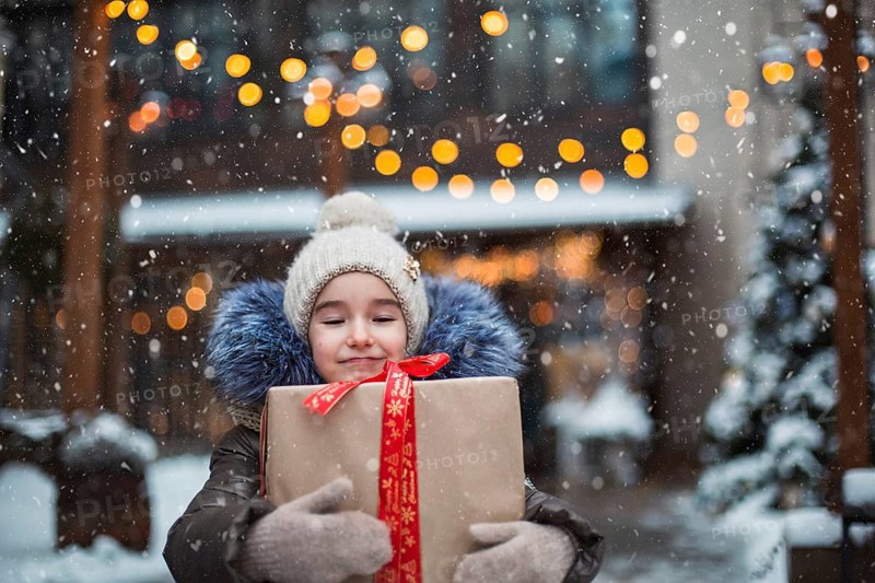 Portrait of joyful girl with a gift box for Christmas on a city street in winter with snow on a festive market with decorations and fairy lights. Warm clothes, knitted hat, scarf and fur. New year