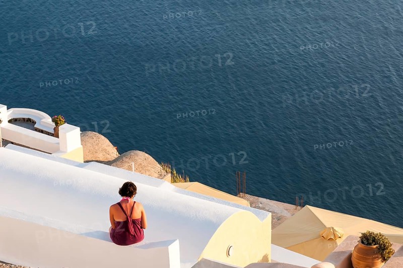Unrecognised woman sitting on the white roof of a house looking at the sea. Santorini island in the Aegean sea, Greece, Europe