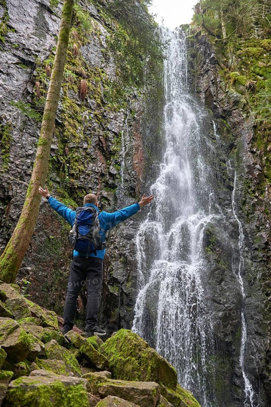Tourist attraction of Germany, falls of Burgbach Waterfall near Schapbach, Black Forest, Baden-Wurttemberg, Germany. Man hiker in blue jacket standing on stone and looks at flow of falling water. Waterfall Landscape and Traveler hands raised enjoying waterfall view Travel Lifestyle adventure concept active vacations into the wild harmony with nature. Perfect view of famous powerful Burgbach Waterfall cascade in sunlight. Dramatic and gorgeous scene. Unique place on earth. Location place Germany, sightseeing Europe. Explore the world's beauty and wildlife. Burgbach Waterfall in gorge of mountains. Amazing beautiful natural waterfall called burgbachwasserfall of river burgbach in black forest nature landscape, a magical place. Germany