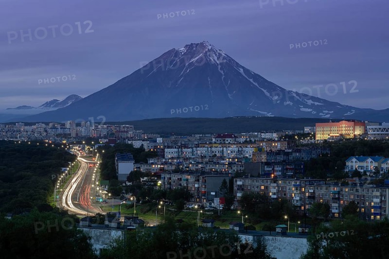 Summer cityscape of Kamchatka Peninsula: night view of urban development of Petropavlovsk-Kamchatsky City, driving automobiles on city asphalt road on background of volcano. Eurasia, Russian Far East