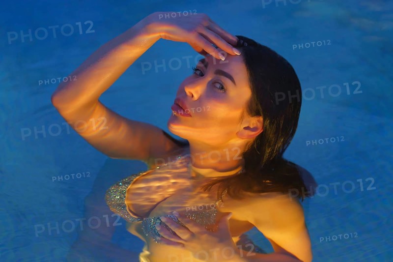 Glamorous brunette in swimsuit swims in geothermal water in pool at spa, looking at camera. Woman's curvy body is illuminated under water by night lights in pool. Soft selective focus on model's eyes