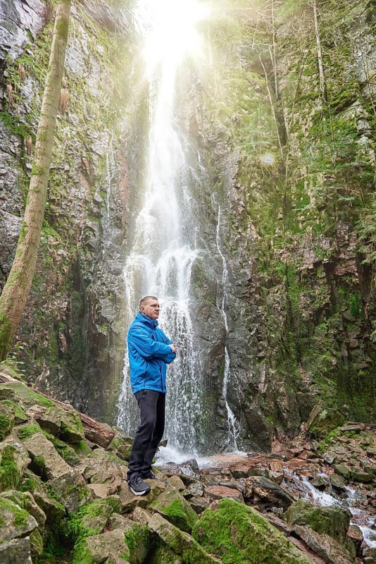 Tourist attraction of Germany, falls of Burgbach Waterfall near Schapbach, Black Forest, Baden-Wurttemberg, Germany. Man hiker in blue jacket standing on stone and looks at flow of falling water. Waterfall Landscape and Traveler hands raised enjoying waterfall view Travel Lifestyle adventure concept active vacations into the wild harmony with nature. Perfect view of famous powerful Burgbach Waterfall cascade in sunlight. Dramatic and gorgeous scene. Unique place on earth. Location place Germany, sightseeing Europe. Explore the world's beauty and wildlife. Burgbach Waterfall in gorge of mountains. Amazing beautiful natural waterfall called burgbachwasserfall of river burgbach in black forest nature landscape, a magical place. Germany