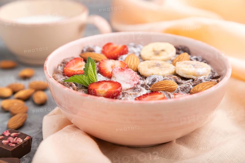 Chocolate cornflakes with milk, strawberry and almonds in ceramic bowl on gray concrete background and orange linen textile. Side view, close up, selective focus