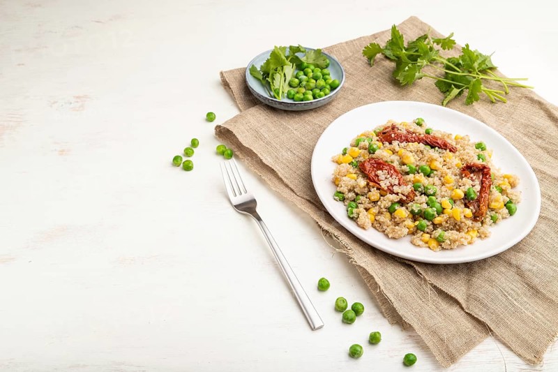 Quinoa porridge with green pea, corn and dried tomatoes on ceramic plate on a white wooden background and linen textile. Side view, copy space