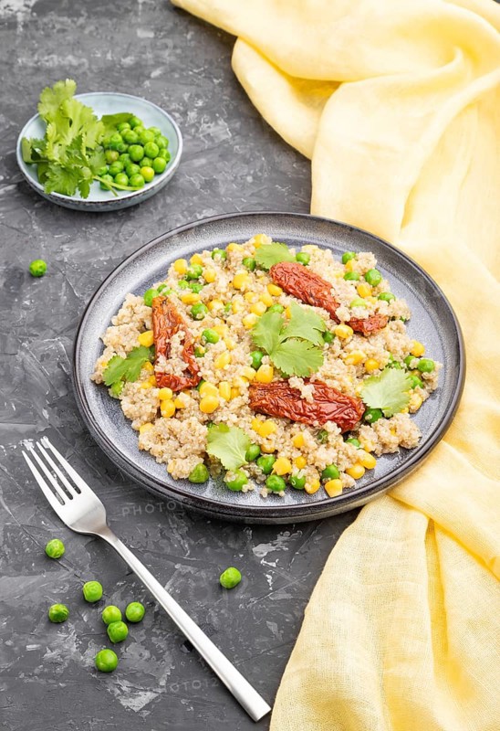 Quinoa porridge with green pea, corn and dried tomatoes on ceramic plate on a gray concrete background and yellow textile. Side view, close up