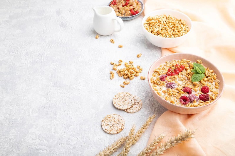 Wheat flakes porridge with milk, raspberry and currant in ceramic bowl on gray concrete background and orange linen textile. Side view, copy space