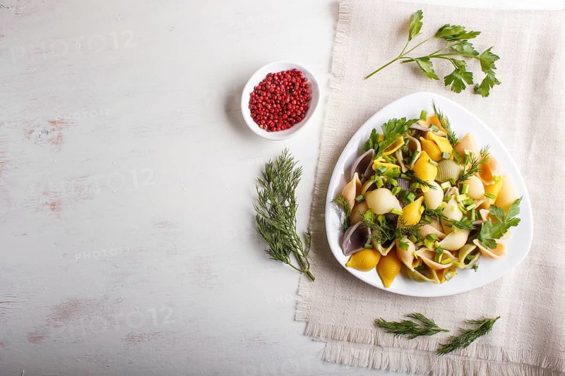 Conchiglie colored pasta with fresh greengrocery on a linen tablecloth on white wooden background. top view, copy space