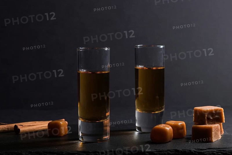 Glass of whiskey with caramel candies on a black stone slate board on black background. Side view, close up, low key concept