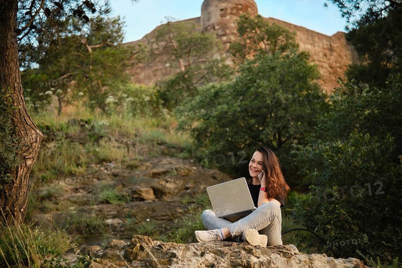 Cheerful woman finds serenity in remote work, seated on rocky terrain with a laptop, surrounded by the lush greenery of a scenic hillside next to ancient Georgian fortress