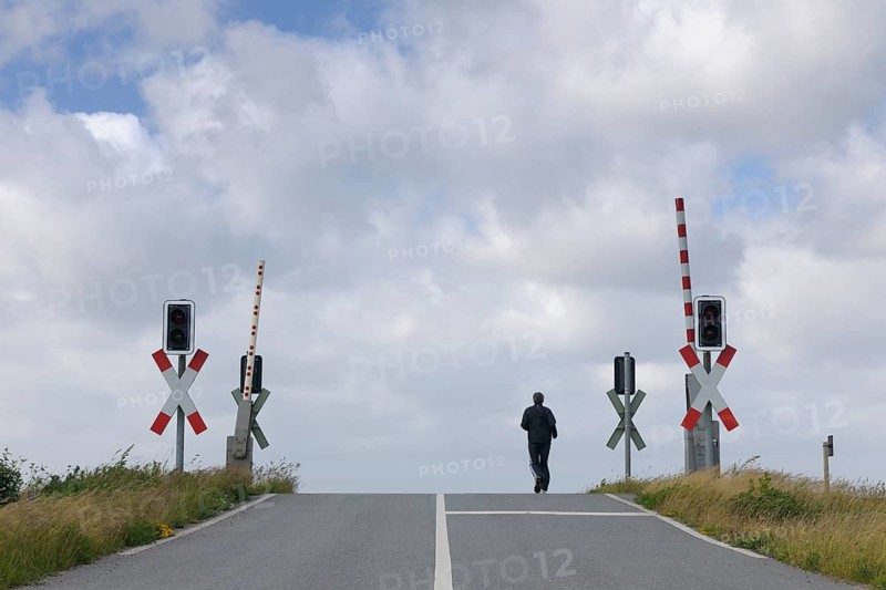 A man walks along an empty road approaching a railway crossing with traffic lights under an overcast sky, Sylt, North Frisian Islands, Hörnum, North Sea, Schleswig Holstein, Germany, Europe
