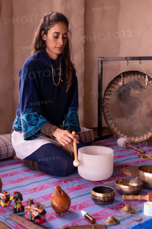 A lovely Latina woman sitting on her ankles, surrounded by sound instruments, engages in meditation, radiating a serene expression as she gently plays a quartz singing bowl