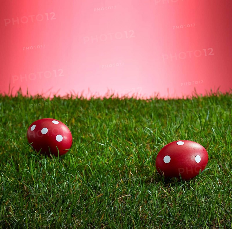 Turf in front of a red background, on it 2 red Easter eggs with white dots, studio shot